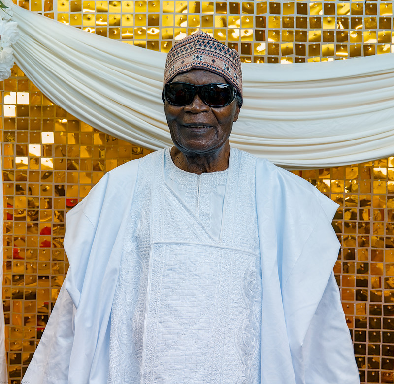 The celebrant in white agbada and cap stands before a gold sequin wall