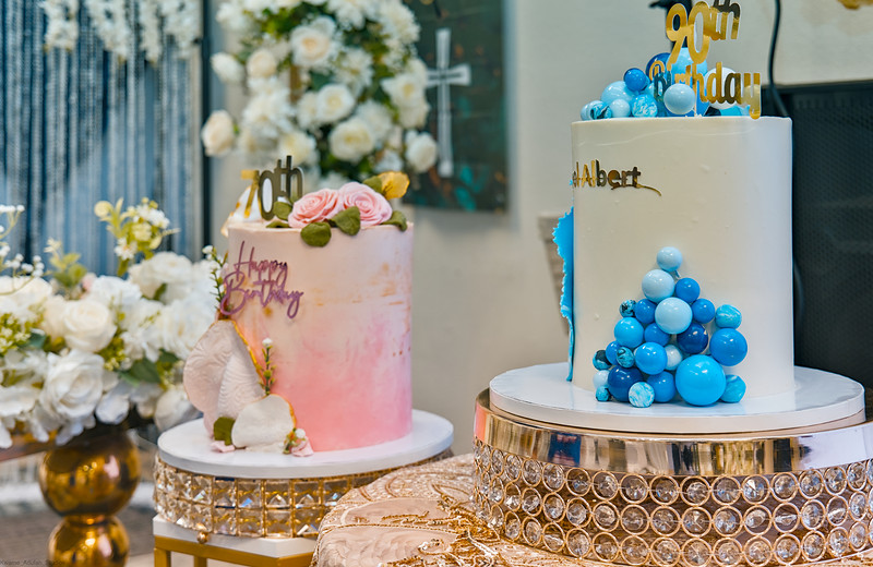 Two birthday cakes, one pink and one blue, on gold stands among white flowers