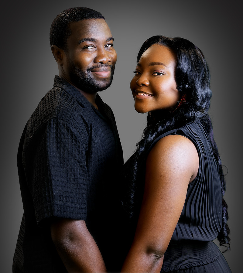 An engaged couple in black, cheek to cheek, smiling at the camera